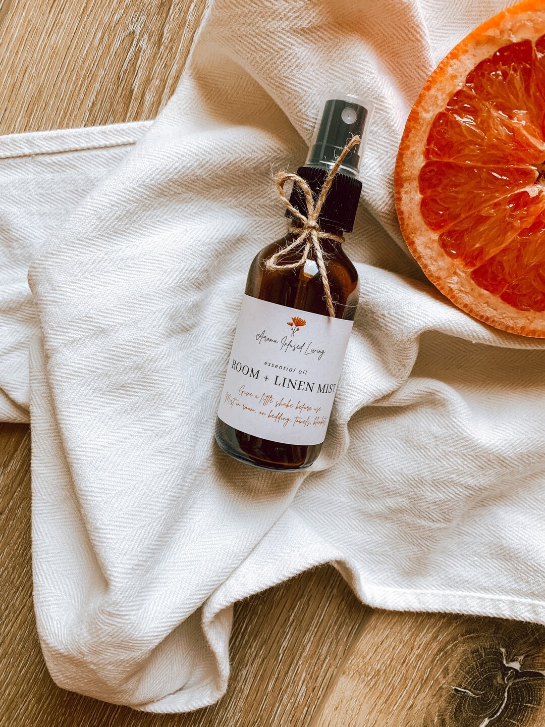 A small amber room and linen mist bottle tied with twine, resting on cream linen beside a halved citrus fruit