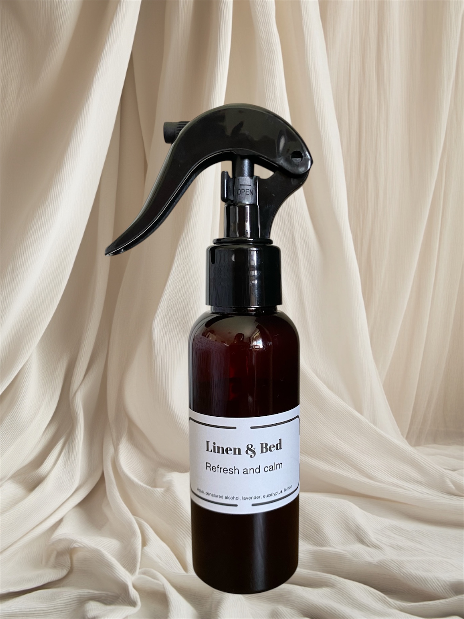 An amber linen spray bottle with a trigger sprayer standing against draped cream fabric