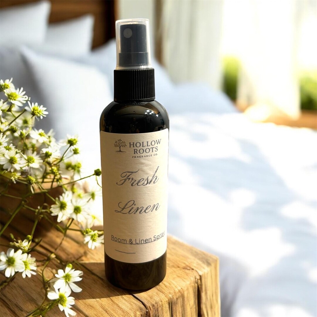 A room and linen spray bottle on a wooden stool beside white daisies, with a softly lit bed behind it